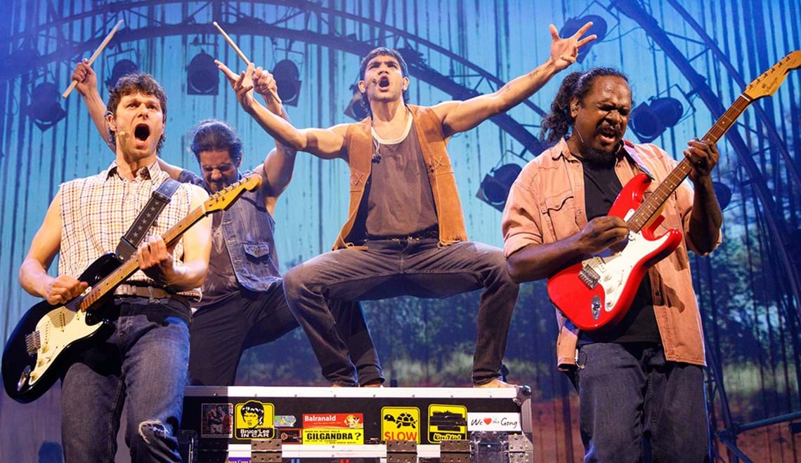 Four people in a band are singing and playing instruments. One person is standing on top of a box with tour stickers, he has his arms out wide as he sings. Two band members are playing guitars and the other is holding drum sticks in the air,