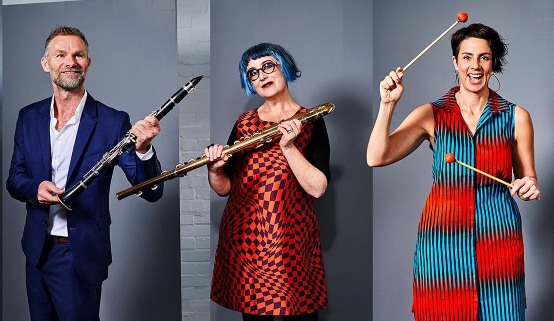 Three people, one man and two women, are holding instruments. The two women wear brightly coloured dresses in shades of orange, while the man wears a navy blue suit. Their expressions and poses are playful.