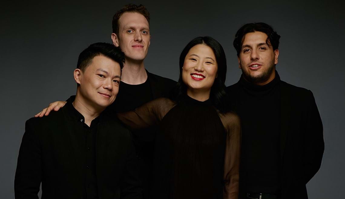 Four musicians stand close together, they are all wearing black. They are slightly smiling and are in front of a dark background