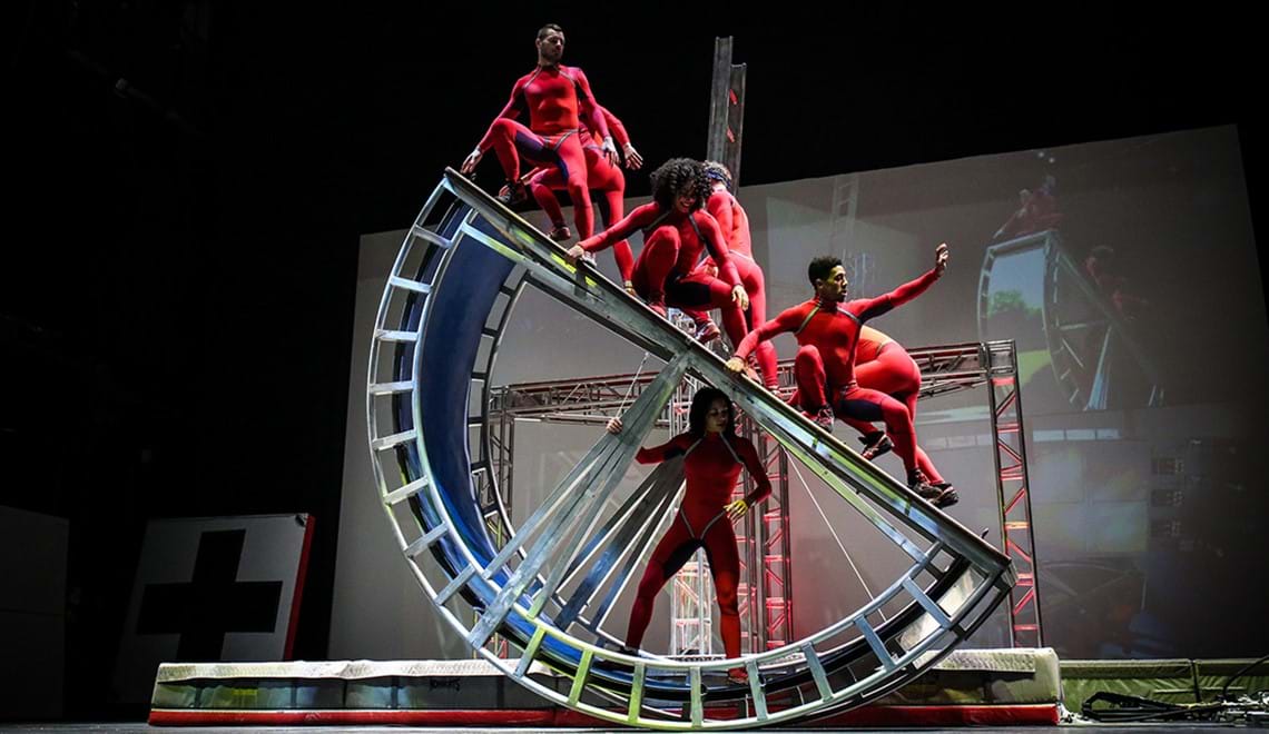 Three performers dressed in red balance on a large 'action machine' - a very large half-wheel.