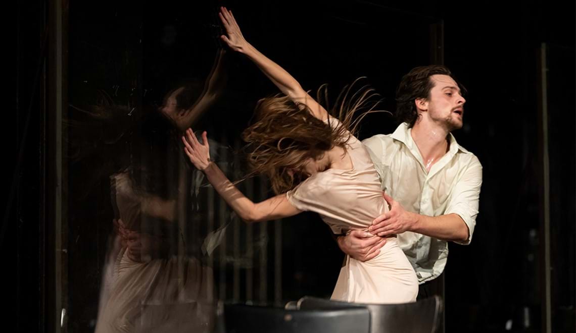 Two dancers in motion. One has a beard and is holding the other around the waist. The other dancer has their arms out and their hair is flung behind them.