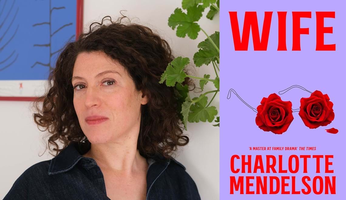 Charlotte Mendelson wears a collared shirt and stands in front of a white wall. Charlotte has curly brown hair.