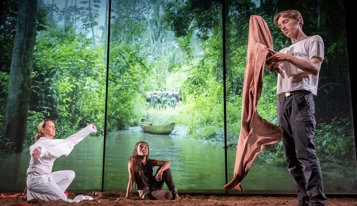 Three actors are in front of a screen - two sit while a third stands while holding a blanket. The screen behind them depicts water and a rainforest with a group of people retreating.