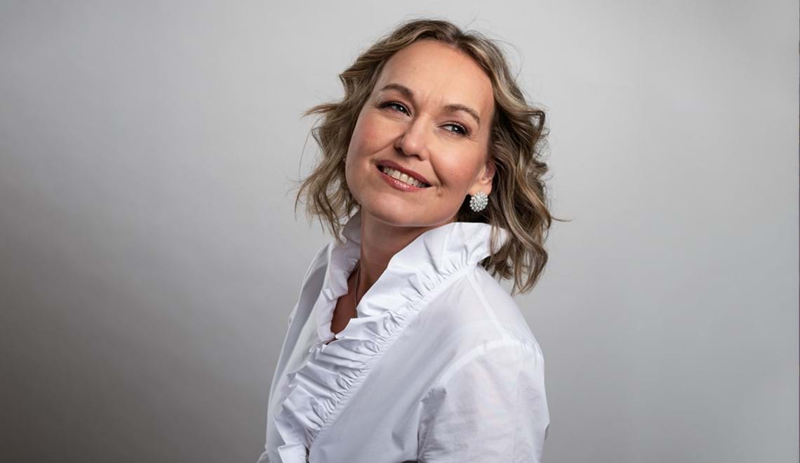 Jenny Carlstedt stands in front of a grey background. She is smiling and looking back over her shoulder. She wears a white shirt that has a ruffle collar and round sparkly earrings. She has curled blonde, shoulder length hair.