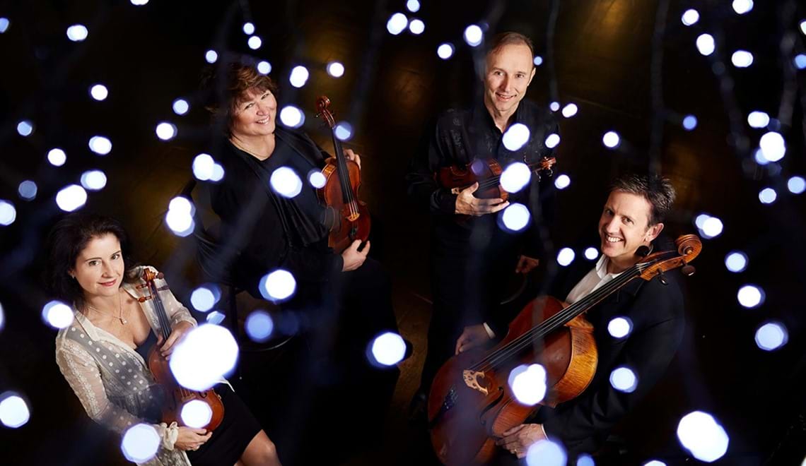 Four people, two men and two women, are wearing black and white and smiling at the camera while holding string instruments.