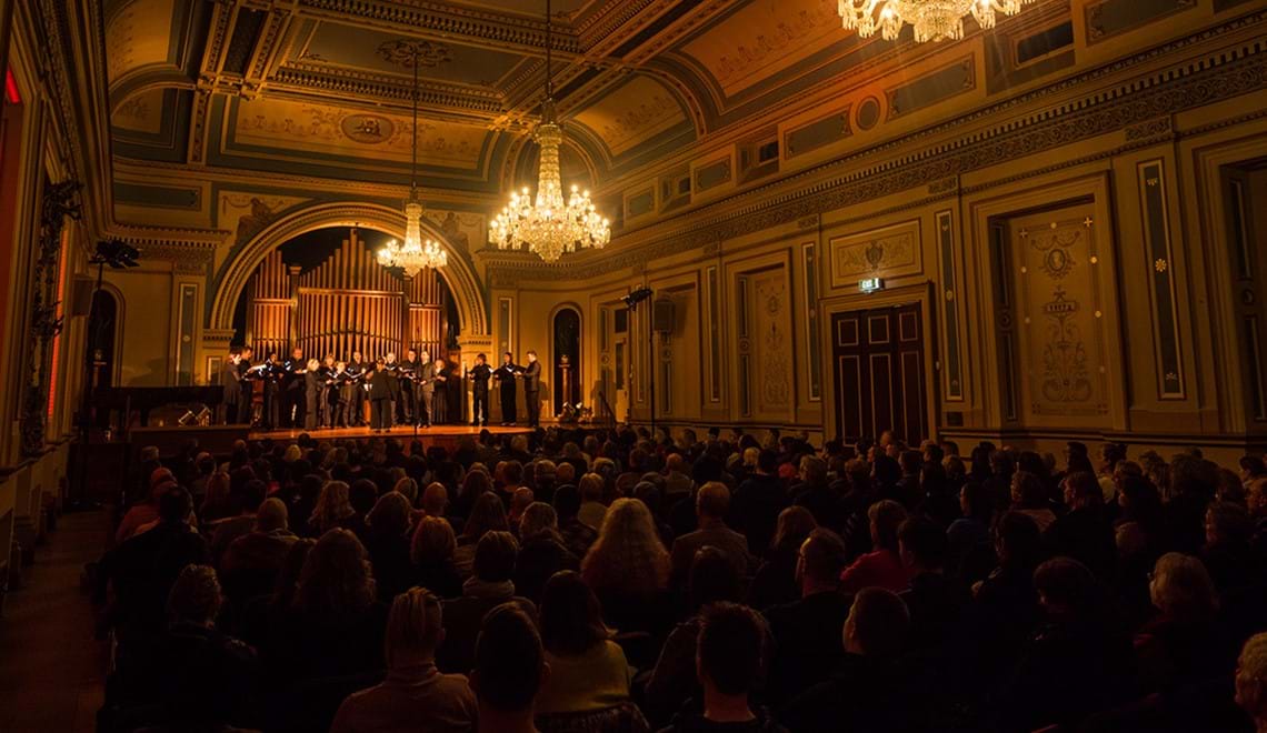A group of singers dressed in black stands on a stage in a dimly lit hall. The heads of the audiences are visible with chandeliers overhead.